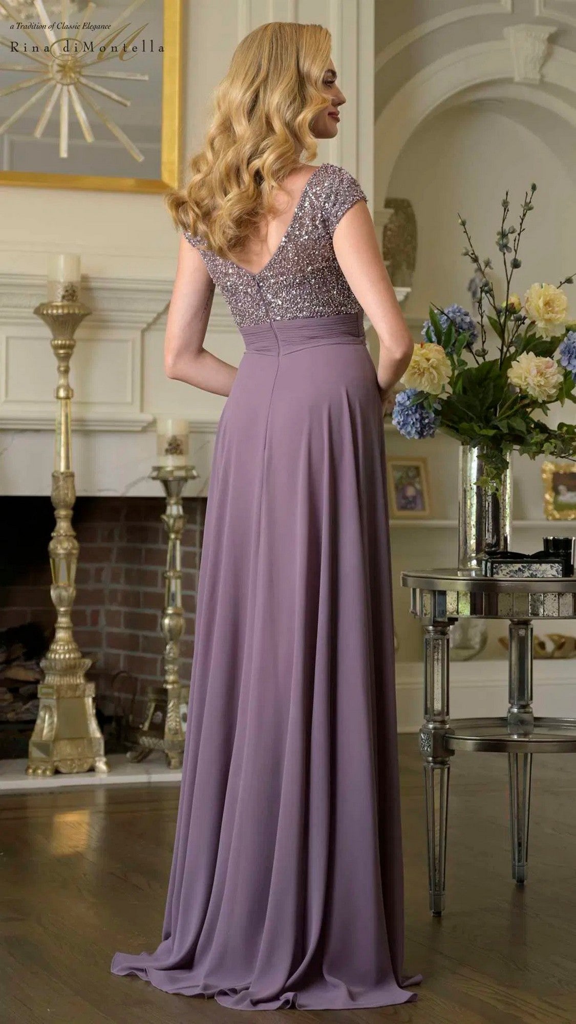 A woman with long blond hair wears the Rina Di Montella RD3419 long formal dress, featuring a beaded bodice and chiffon flowy skirt, as she stands in an elegant living room facing away from the camera.